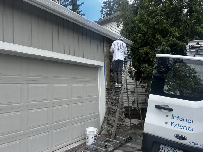A painter stands on a ladder painting a house exterior next to a garage, with a Hendricks "Interior Exterior" van parked nearby. Scene captured in May 2025; location not specified as Portland or Pearl Painters.