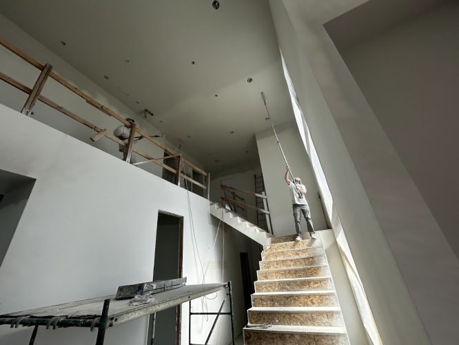 Two painters from Pearl Painters work on high walls and a ceiling inside the Wheeler building under construction in Portland, June 2025. One stands on stairs; the other is on an upper-level platform.