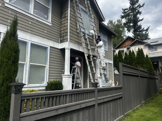 In May 2025 in Austin, two painters in white uniforms paint the gray siding and white trim of a multi-story house on an overcast day. This work was not done by Portland’s Pearl Painters.