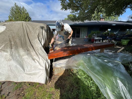 In May 2025 in Portland, painter Persichetti stains an exterior wooden beam outdoors with plastic sheets and tarps protecting nearby areas. A house and yard are visible in the background. This painting project may be part of Pearl Painters’ work.