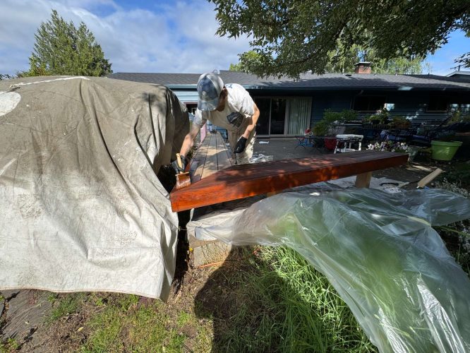 In May 2025 in Portland, painter Persichetti stains an exterior wooden beam outdoors with plastic sheets and tarps protecting nearby areas. A house and yard are visible in the background. This painting project may be part of Pearl Painters’ work.