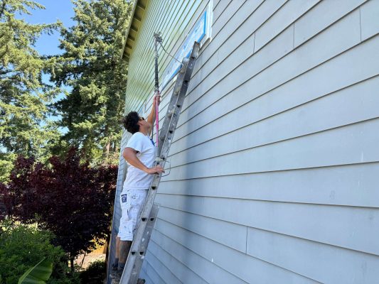 In May 2025, a painter from Pearl Painters uses a roller and ladder to paint the upper exterior wall of a house in Portland light green.