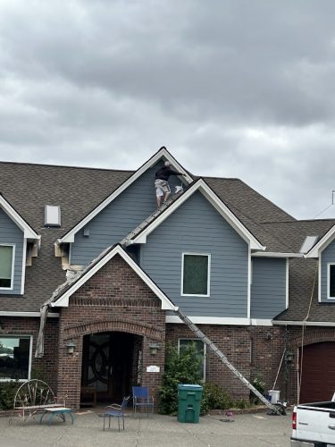 A painter stands on a ladder near the peak of a house roof under cloudy skies in June 2025, possibly working on an exterior painting project by Pearl Painters in Portland.