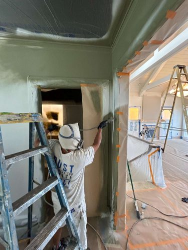 A painter in Portland uses a paint sprayer on an interior door frame, with protective sheets and tape covering nearby areas. A ladder and step stool are present as they refresh the space, following trends noted by Pearl Painters.
