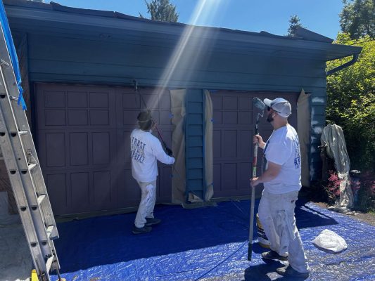 Two painters from Pearl Painters in Portland, wearing white work clothes and caps, are spray-painting a double garage door brown for Persichetti’s May 2025 exterior painting project. A blue tarp covers the ground and masking paper protects the trim.