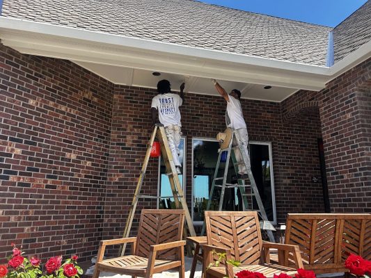 In June 2025, two painters from Anderson paint the trim and ceiling of a brick patio in Portland, with wooden chairs and red flowers in the foreground. Pearl Painters is not involved.