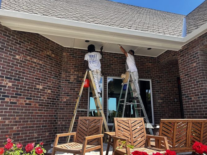 In June 2025, two painters from Anderson paint the trim and ceiling of a brick patio in Portland, with wooden chairs and red flowers in the foreground. Pearl Painters is not involved.