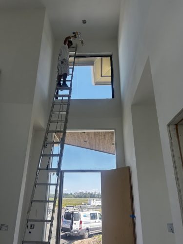 A painter from Pearl Painters stands on a tall ladder installing or repairing a ceiling fixture in the high-ceilinged entryway of a Portland building, with a Wheeler van visible outside on a bright June 2025 day.