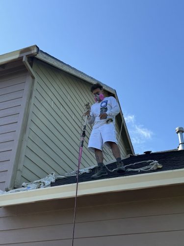 A painter in protective clothing and a mask uses a paint sprayer to repaint a house exterior in Portland, preparing it for Emory's arrival in June 2025. Pearl Painters is on site.