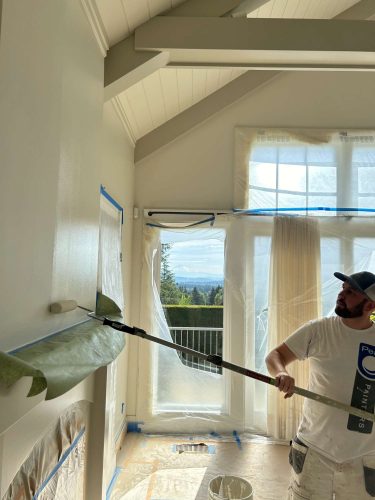 A painter from Pearl Painters in Portland uses a roller to paint a high interior wall, with plastic and painter’s tape protecting surfaces—showcasing a fresh interior update by Duffy in May 2025.