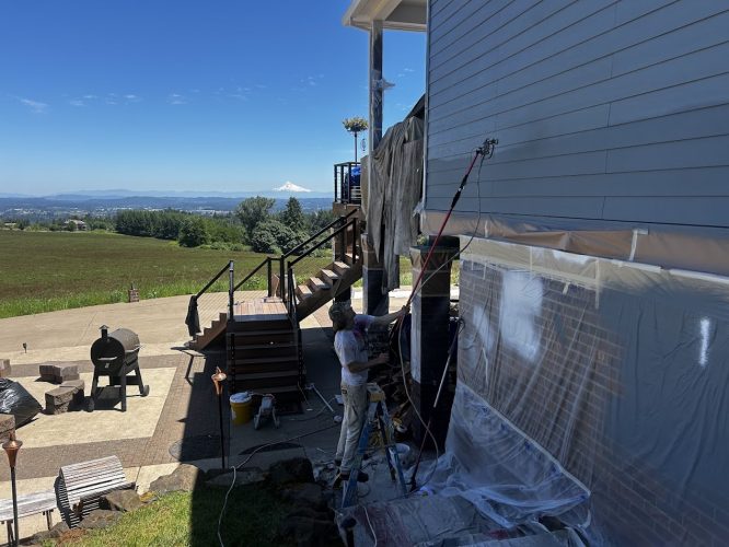 A painter from Pearl Painters uses a sprayer to paint the exterior siding of an Anderson home, with plastic sheets protecting nearby surfaces. The work takes place near Portland, with a scenic landscape and mountain visible in the background on a sunny June 2025 day.