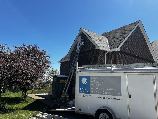 In June 2025, a painter from Pearl Painters works on the exterior of a brick house in Portland using a tall ladder, with a company trailer nearby.