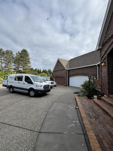 Two white vans belonging to Pearl Painters are parked in the driveway of a large brick house with three garage doors in Portland. The sky is cloudy, trees are visible, and “Anderson” appears on the exterior. Photo taken June 2025.