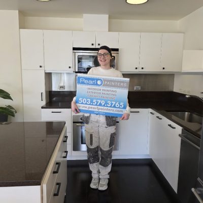 Painter in paint-splattered clothes holds a Pearl Painters sign for painting services in a modern Portland kitchen.
