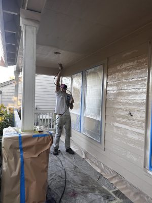 Painter in protective gear spray painting porch ceiling; windows/items covered for protection. Pearl Painters, Portland.