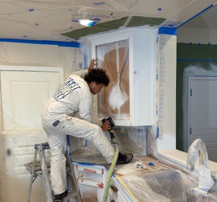 A painter from Pearl Painters in Portland sprays paint on a kitchen cabinet, with plastic sheeting protecting the area.