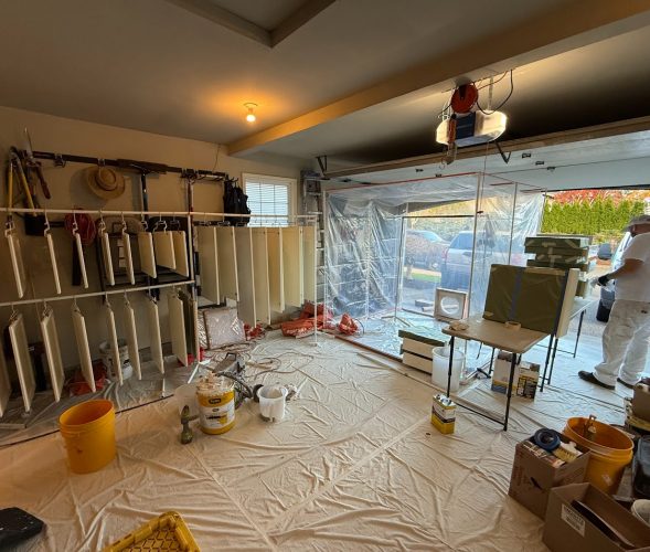A painter in Portland dries cabinet doors on racks in a garage prepped for painting by Pearl Painters. Tools and paint cans visible.
