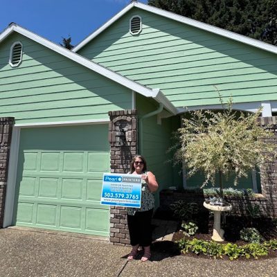 A woman stands in front of a green house with brick trim in Portland, holding a Pearl Painters painting company sign.