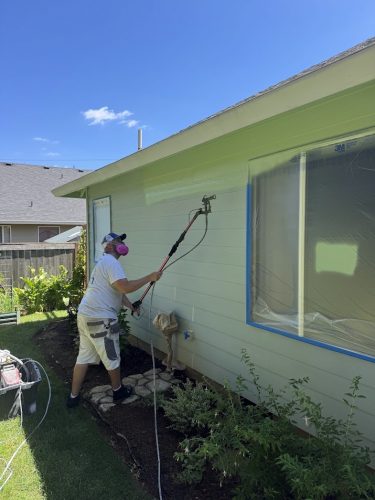 Painter in protective gear paints a house exterior light green with a sprayer; Pearl Painters, Portland.