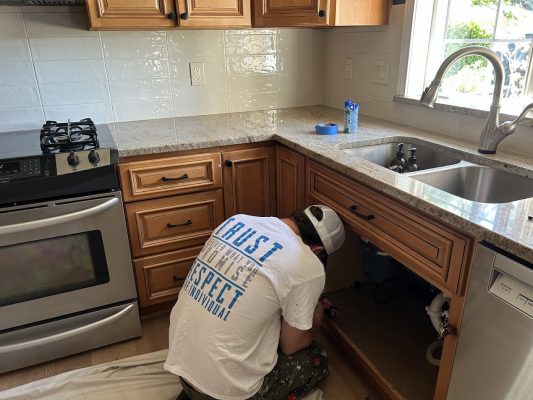 A person repairs plumbing under a kitchen sink with brown cabinets. No painter or painting, no Pearl Painters, location not Portland.