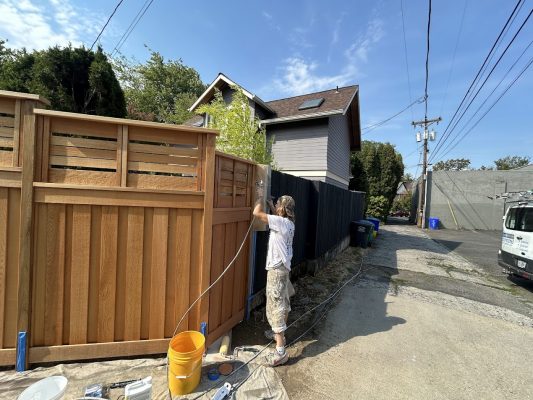 A painter from Pearl Painters uses a spray gun to paint a wooden fence on a sunny day in a Portland residential alley.
