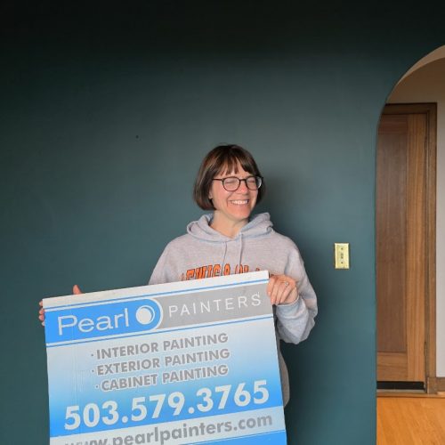 Person in glasses holds Pearl Painters sign for painting services, standing by a dark teal wall in Portland.