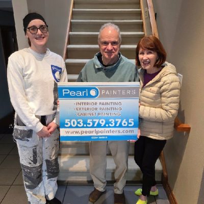 Three people indoors at a staircase hold a Pearl Painters sign showing painter services in Portland and contact info.