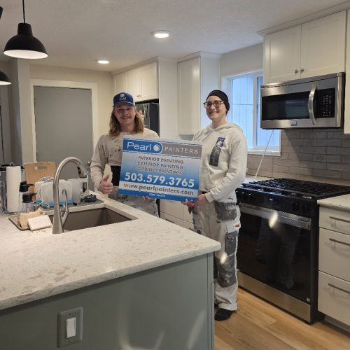 Two painters in work clothes hold a Pearl Painters sign in a modern Portland kitchen. Sign lists contact info and services.