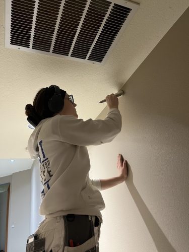 A painter in a white hoodie paints a beige wall edge near an air vent; likely for Pearl Painters in Portland.