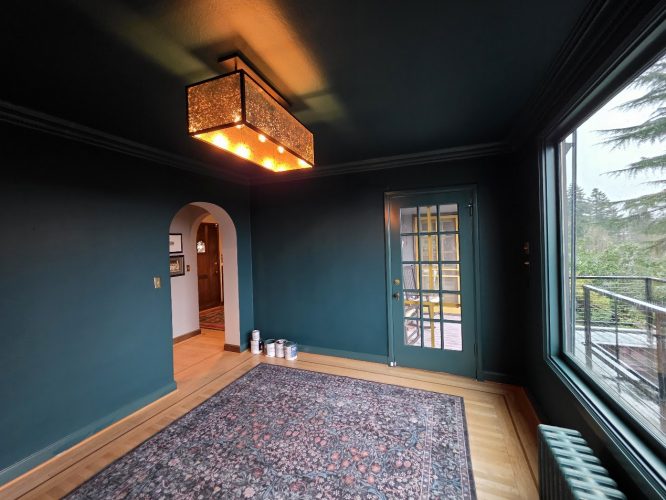 A Portland room painted dark teal, with a patterned rug, ceiling light, paint cans—painting by Pearl Painters. Balcony door visible.