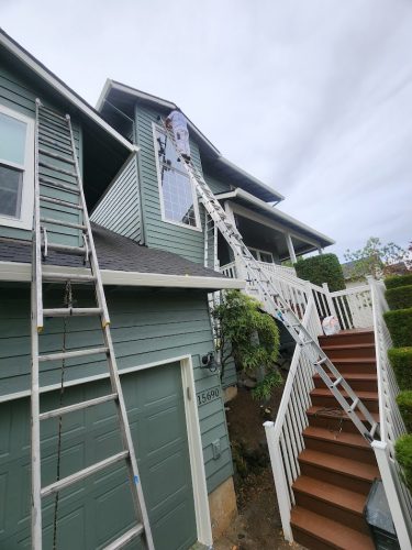A painter on a tall ladder paints a two-story house’s exterior near a window above the garage; overcast day, Pearl Painters, Portland.