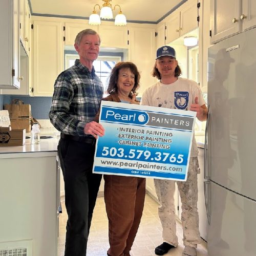 Three people in a kitchen in Portland hold a Pearl Painters sign for painting services; cabinets and appliances are visible.