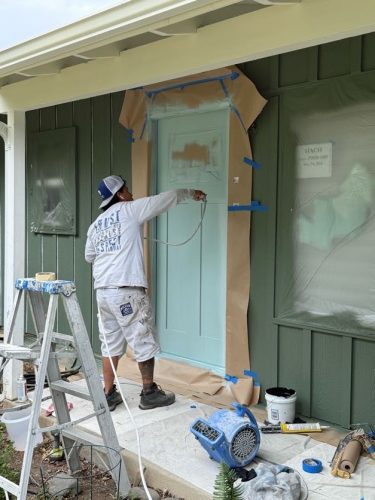 A painter uses a paint sprayer to paint a door light blue, with tape and paper masking the area; tools and supplies are nearby. Pearl Painters, Portland.