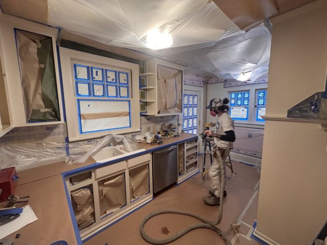 A painter in protective gear sprays cabinets in a plastic-covered room. Painting by Pearl Painters, Portland.