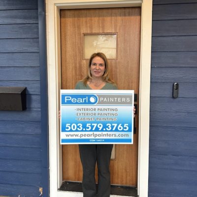 Woman in front of wooden door holds Pearl Painters sign, advertising painting services in Portland with contact info.