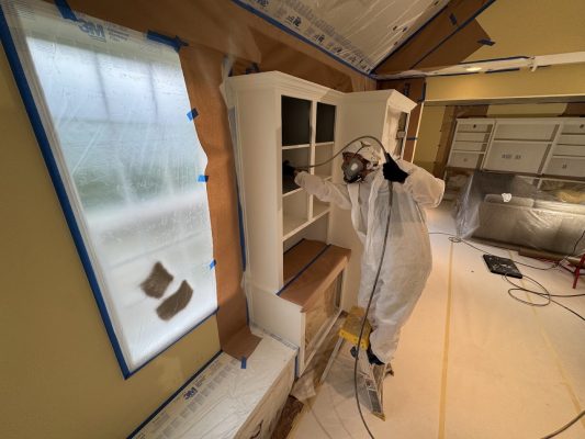 A painter in protective gear stands on a ladder, ready to paint cabinets in a prepped room, likely for Portland’s Pearl Painters.