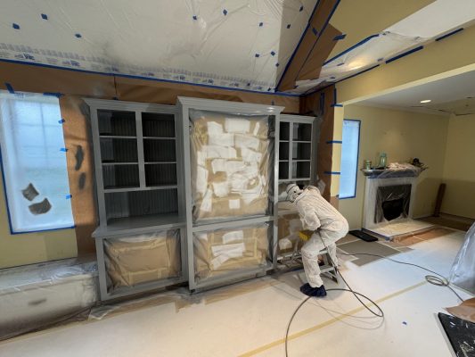 A painter in protective gear uses a sprayer to paint cabinets in a taped-off room. Pearl Painters, Portland.