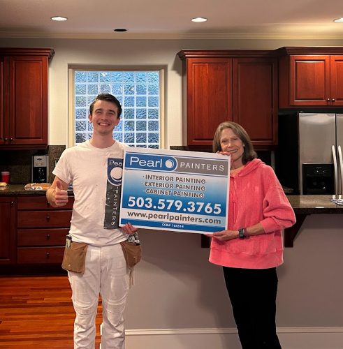 A painter and woman in a kitchen hold a Pearl Painters sign for painting services in Portland, including contact info.