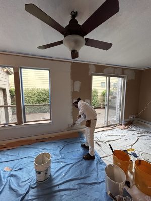 A painter in white work clothes paints a wall near large windows in Portland. Paint tools and Pearl Painters buckets are visible.