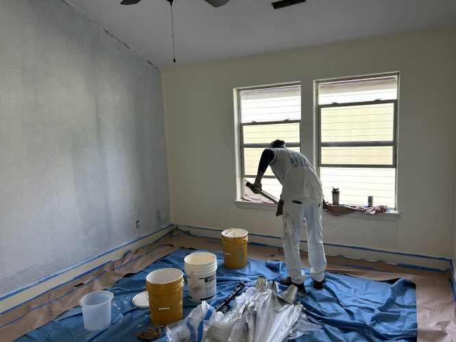 A painter from Pearl Painters works in a room, painting a wall. Tarps cover the floor and supplies are scattered around.