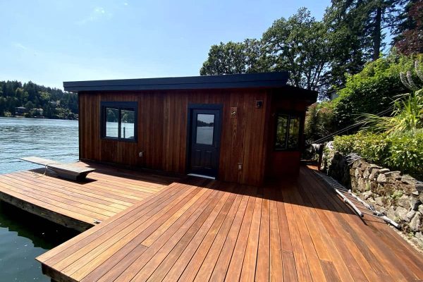A wooden houseboat with large windows and a black door is situated on a lakeside deck, surrounded by trees and water, with deck and house fully refinished and stained by Pearl Painters in Portland