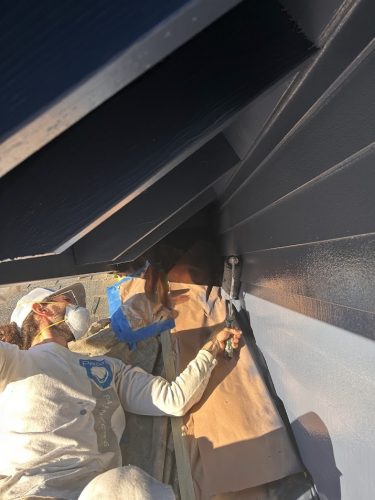 A painter in work clothes and mask paints house siding dark blue with a brush, paper protecting the ground; Pearl Painters, Portland.