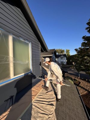 A painter in work clothes on a roof uses a roller to paint a house exterior; window covered. Pearl Painters, Portland.