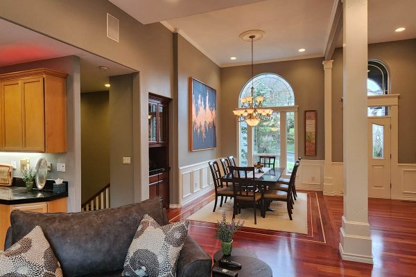 Elegant dining room with a large table, hardwood floors, a chandelier, and adjacent living room with a gray sofa. decor includes large landscape paintings and warm lighting.