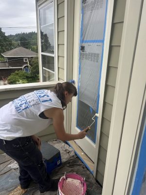 A painter in a sleeveless shirt is painting a door frame white on an outdoor balcony, with windows protected by plastic and painter’s tape—work possibly by Pearl Painters in Portland.
