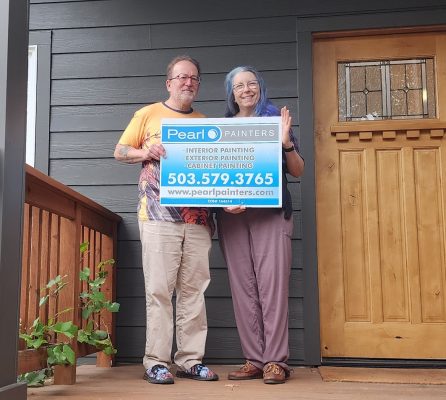 Two people on a porch in Portland hold a Pearl Painters sign with painting services and phone number by a wooden door.