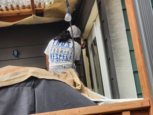 A painter in a cap and blue-lettered shirt paints door trim on a covered patio. Scene is likely Portland, Pearl Painters.