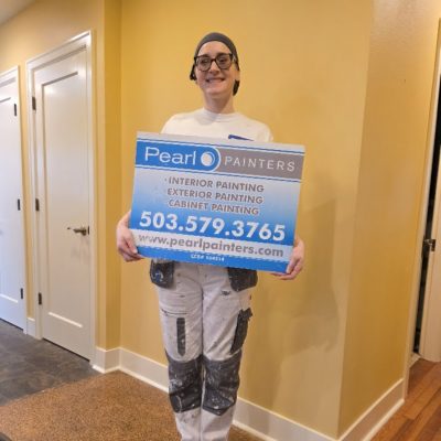 Painter in work clothes holds Pearl Painters sign for painting services in Portland, showing phone number and website inside.