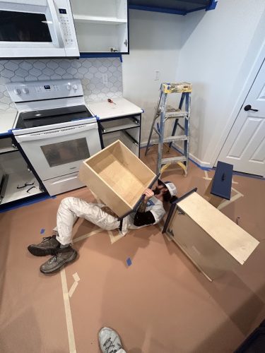 A person lies on the floor fixing a kitchen drawer near a stove; ladder and tools visible. Painter, painting, Pearl Painters, Portland.