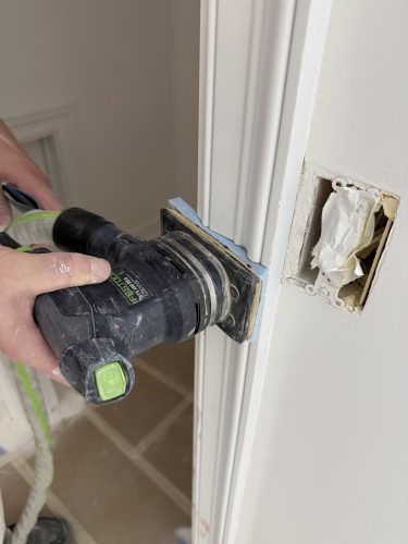 A painter in Portland uses an electric sander on a white door frame near an uncovered outlet, prepping for Pearl Painters.